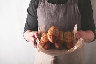 Руки мужчины, держащего горячий запеченный сэндвич на багетном хлебе с ветчиной, беконом, овощами и сыром на пергаменте мужчина в черном фартуке на фоне домашней кухни с пекарней макет