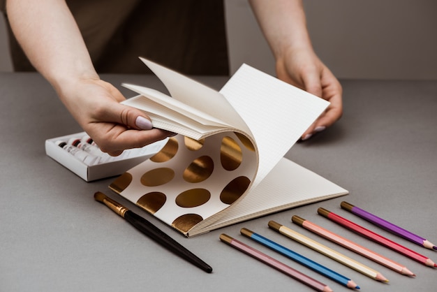 Free photo hands opening sketchbook with oil paints and pencils on grey table