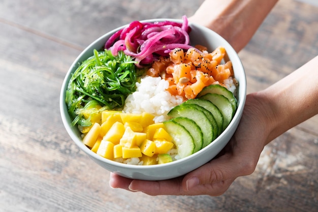 Foto gratuita mani che mostrano una poke bowl con riso salmonecetriolomangoonionwakame insalata semi di papavero e semi di girasoli su legno tablexa