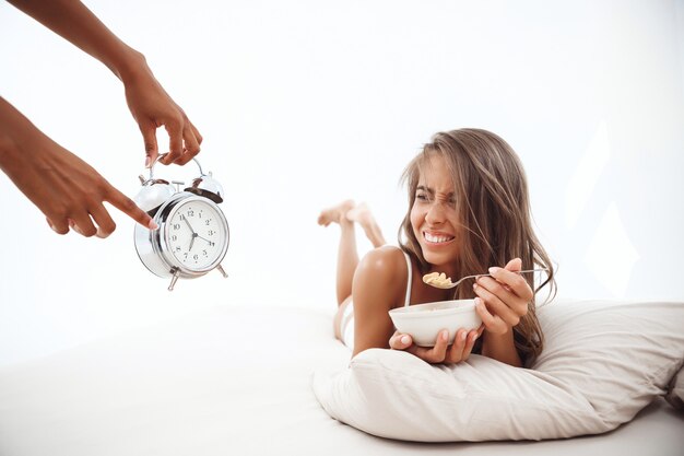 ベッドに横になっている美しい女性に目覚まし時計で時間を示す手
