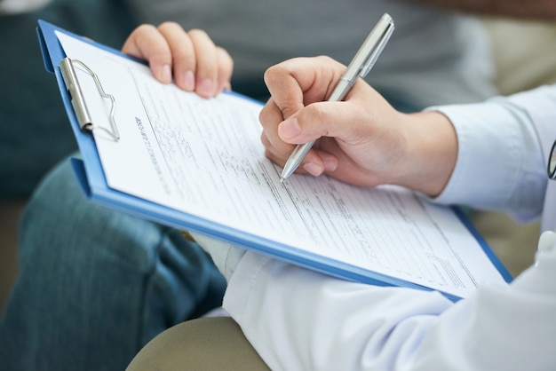 Hands of unrecognizable female doctor filling in form on clipboard