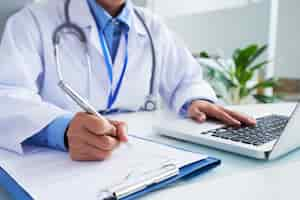 Free photo hands of unrecognizable female doctor writing on form and typing on laptop keyboard