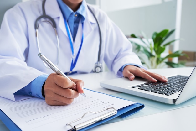 Hands of unrecognizable female doctor writing on form and typing on laptop keyboard