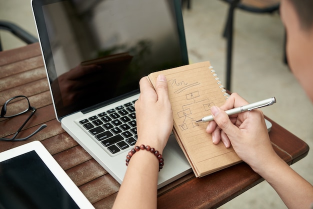 Hands of unrecognizable man sitting at table with laptop and drawing diagram in notebook