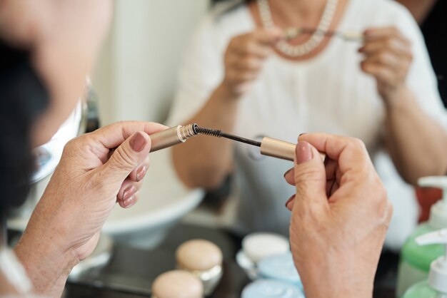 Hands of unrecognizable senior lady holding mascara in front of mirror
