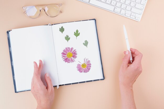 Hands with pen near notebook with dry flowers, keyboard and eyeglasses