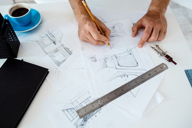 Hands of young businessman holding pencil and drawing sketch at table. White modern office interior 