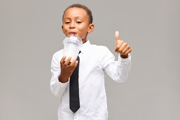 handsome adorable dark skinned male pupil wearing white shirt and black tie making thumbs up gesture while sipping healthy milkshake on lunch break at school, having joyful look