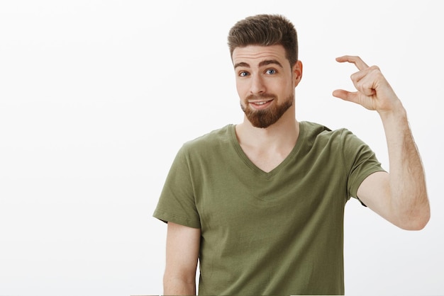 Handsome adult bearded guy in olive tshirt shaping tiny or little thing and looking questioned at front with silly smile as if asking question against white wall