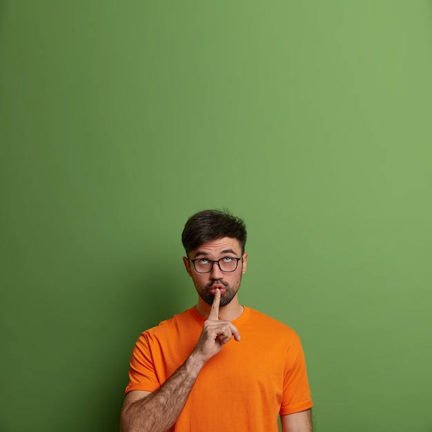 Free photo handsome adult man asks keep quiet, puts finger on mouth, concentrated above, tells secret information, makes hush gesture, dressed in casual orange t shirt, isolated on green wall, copy space up