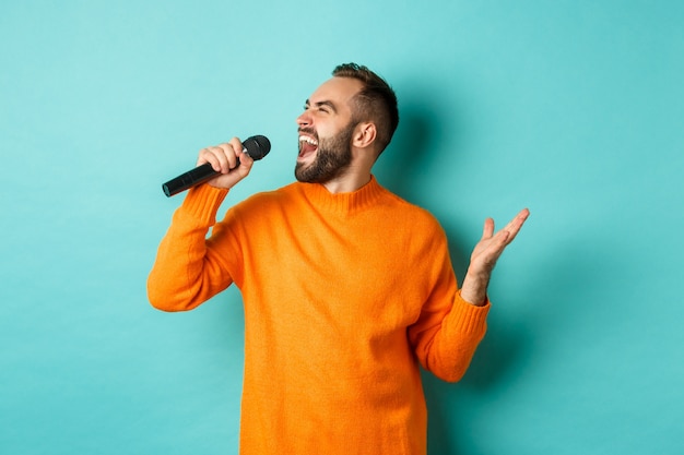 Handsome adult man perform song, singing into microphone, standing against turquoise wall