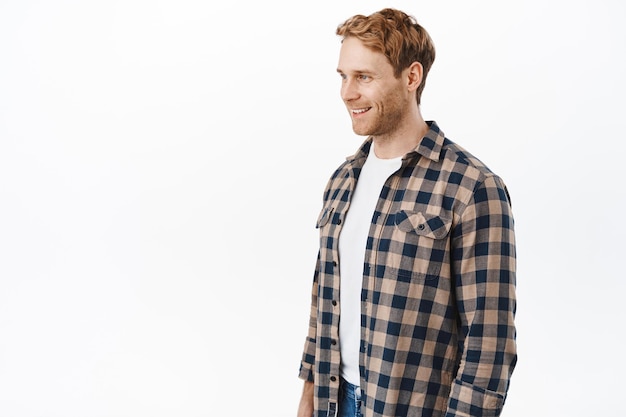 Handsome adult redhead guy with bristle and blue eyes, standing half-turned and looking left at copyspace advertisement, smiling as if talking to someone, standing over white background