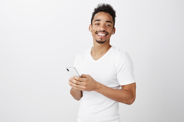 Красивый афро-американский парень слушает музыку в беспроводных наушниках и держит мобильный телефон, довольно улыбаясь
