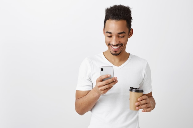 Handsome african american guy in white t-shirt chatting, texting and drinking coffee, looking at mobile phone screen