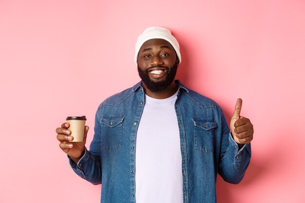 Красивый афро-американский битник мужчина показывает палец вверх, пьет кофе и рекомендует кафе, стоя на розовом фоне.