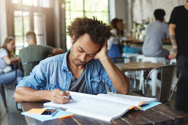 Handsome African American male student feeling tired and stressed as he has to redo homework, trying to focus on task and find where he did mistake, staring at copybook with concentrated look