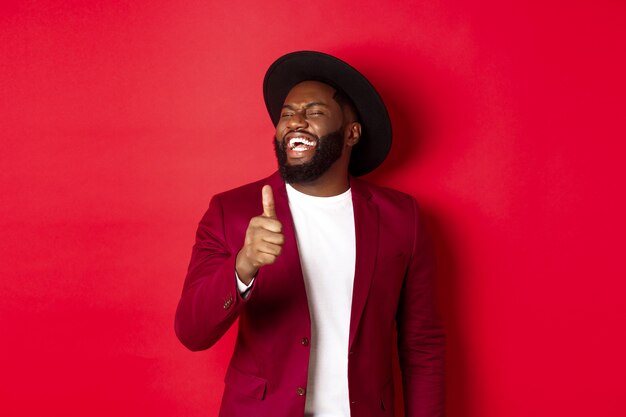 Handsome african american man having fun, showing thumb up and laughing from good joke, standing against red background.