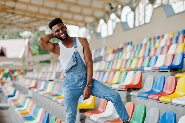 Handsome african american man at jeans overalls posed on colored chairs at stadium Fashionable black man portrait