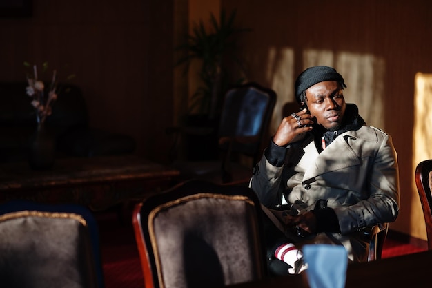 Free photo handsome african american man posing inside room with sunlight shadows in black hat and beige coat speaking on mobile phone