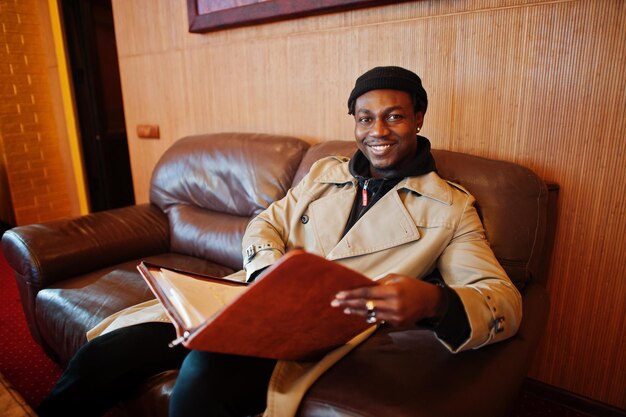 Free photo handsome african american man posing inside room with sunlight shadows in black hat and beige coat