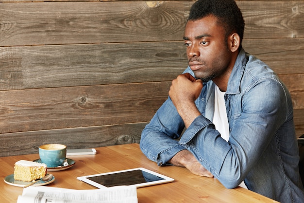 Bello blogger africano in giacca di jeans con uno sguardo pensieroso, toccandosi il mento mentre pensa al suo nuovo post, seduto al tavolino del bar con tazza, torta, giornale e touch pad con schermo vuoto