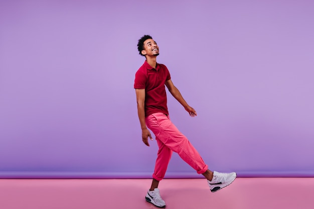 Handsome african guy in trendy pants looking up with smile. Indoor photo of interested black male model dancing.