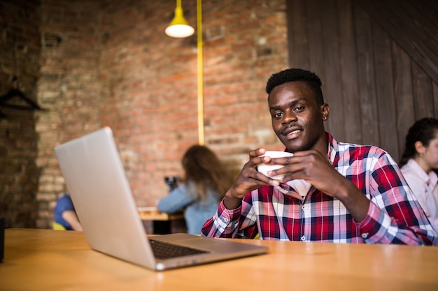 Handsome Afro American man in casual clothes holding a cup of coffee and using laptop.