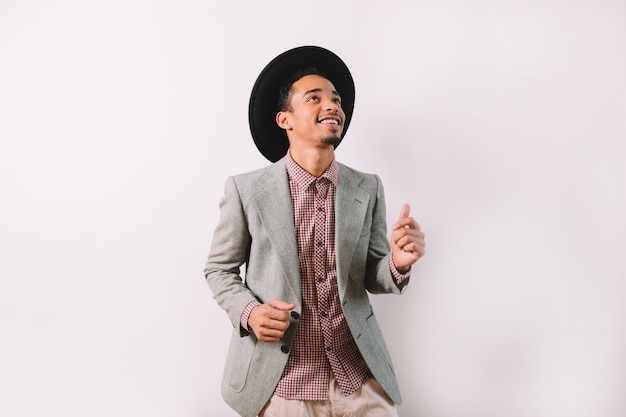 Handsome afro-american man dressed grey jacket and black hat dances with perfect smile on gray