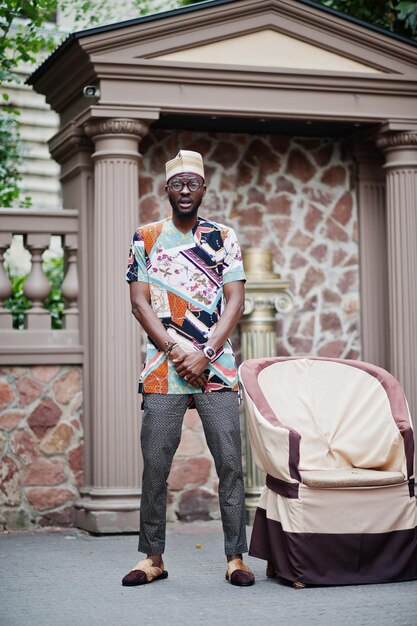 Handsome afro american man wearing traditional clothes cap and eyeglasses in modern city standing near chair