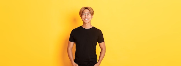 Handsome asian blond guy in glasses standing in black clothes and smiling pleased yellow background