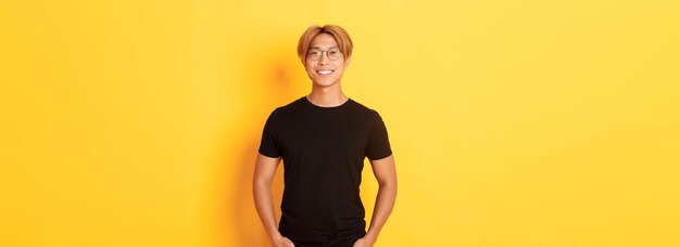 Handsome asian blond guy in glasses standing in black clothes and smiling pleased yellow background