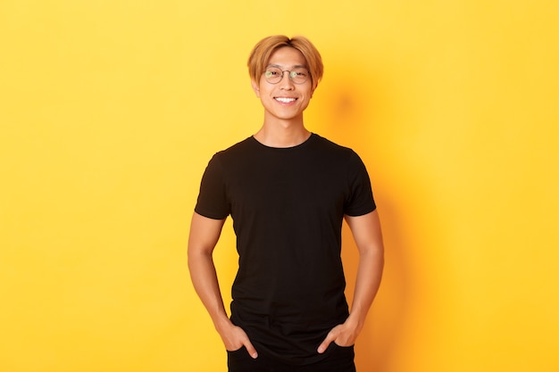 Handsome asian blond guy in glasses standing in black clothes and smiling pleased, yellow wall