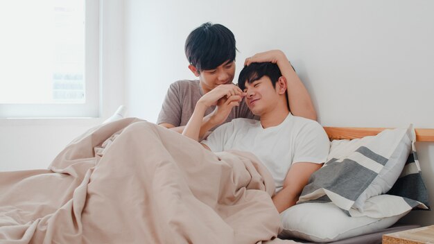 Handsome Asian gay couple talking on bed at home. Young Asian LGBTQ+ guy happy relax rest together spend romantic time after wake up in bedroom at modern house in the morning .