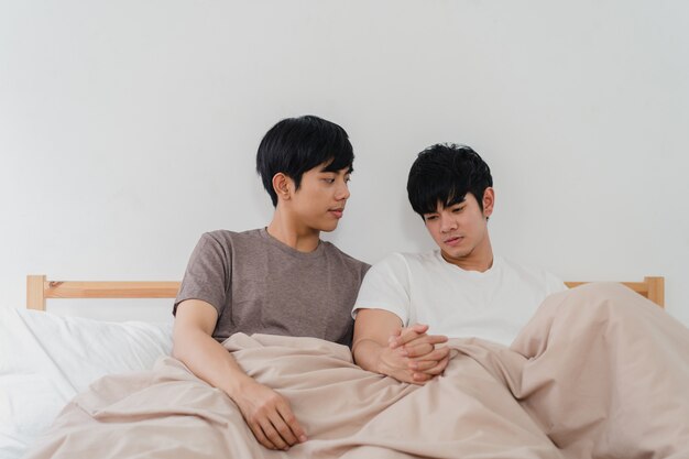 Handsome Asian gay couple talking on bed at home. Young Asian LGBTQ+ guy happy relax rest together spend romantic time after wake up in bedroom at modern house in the morning .