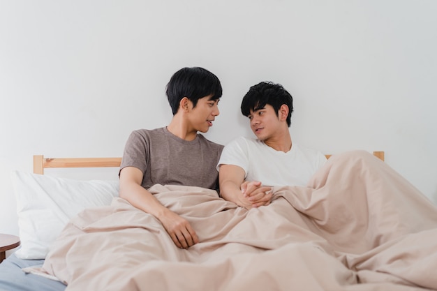 Handsome Asian gay couple talking on bed at home. Young Asian LGBTQ+ guy happy relax rest together spend romantic time after wake up in bedroom at modern house in the morning .