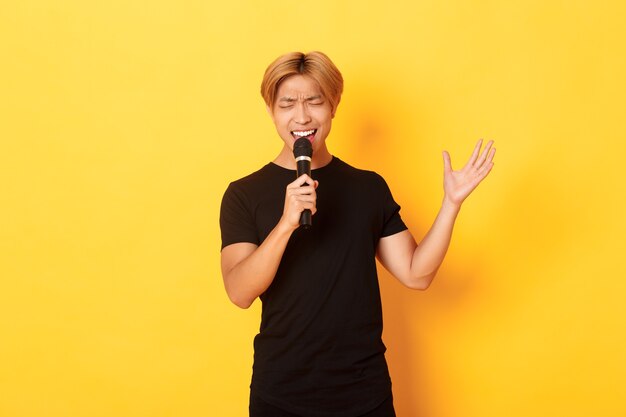Handsome asian male singer, korean guy singing song at karaoke in microphone with passion, standing over yellow wall