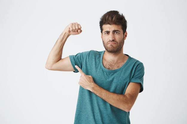 Handsome athletic italian unshaven guy in blue shirt, with concentrated expression showing his muscles after month of training in gym.