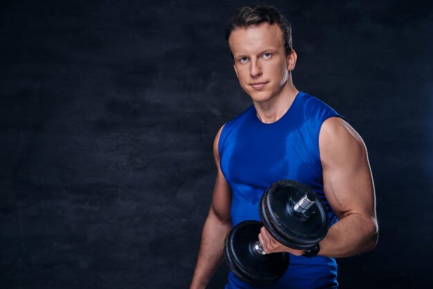 Handsome, athletic male in a blue sportswear holds the dumbbell over dark vignette background.