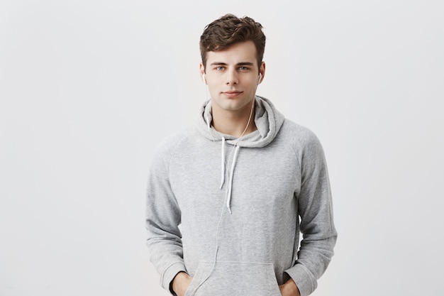 Handsome attractive European man in gray hoodie, with hands in pockets, look pleased , has good mood as comes home after work. Good-looking male student poses .