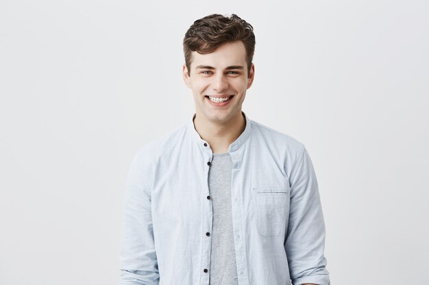 Handsome attractive young man dressed in light blue shirt over t-shirt with dark hair and appealing blue eyes looknig , smiling broadly, demonstrating white teeth, being happy and pleased.