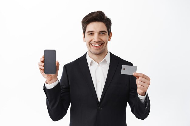 Handsome bank clerk showing smartphone screen and plastic credit card, smiling confident, standing against white background in black suit