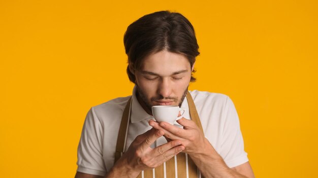 新鮮なコーヒーを嗅ぐエプロンのハンサムなバリスタは彼の仕事が大好きですカラフルな背景の上においしいコーヒーを楽しんでいる若いひげを生やした男