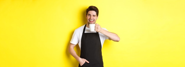 Free photo handsome barista drinking cup of coffee and winking inviting to cafe standing over yellow background