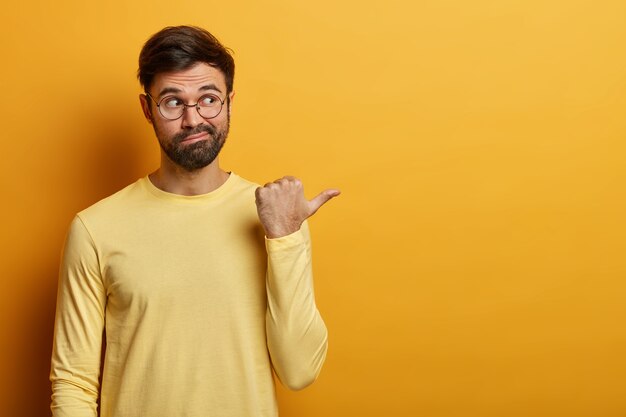 Handsome bearded adult man points thumb aside, shows banner or advertisement, tells to visit online store, dressed casually, isolated on yellow wall, found what he need. Monochrome shot.