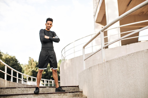 Handsome bearded brunette darkskinned man in black shorts and longsleeved tshirt looks into camera and crosses arms Guy poses on stairs outside