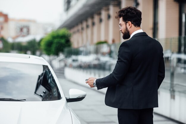 Handsome bearded businessman in black suit entering his car while standing outdoors on the streets of the city near the modern office center