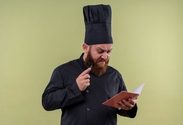 A handsome bearded chef man in black uniform holding pencil while looking at the notebook on a green wall