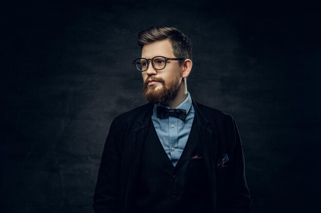 Handsome bearded elegant male in eyeglasses dressed in a dark blue suit with bow tie over grey vignette background.