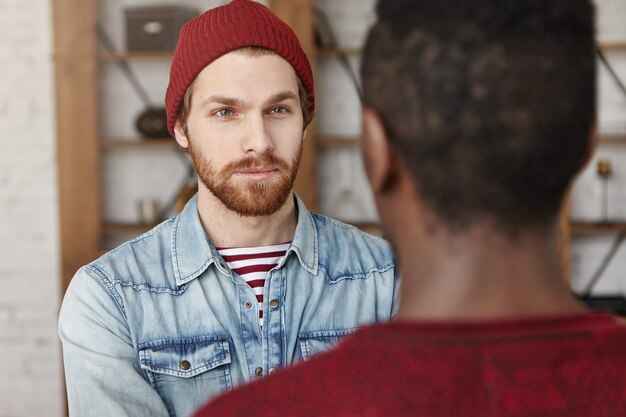 Handsome bearded European hipster wearing hat and denim jacket having serious conversation