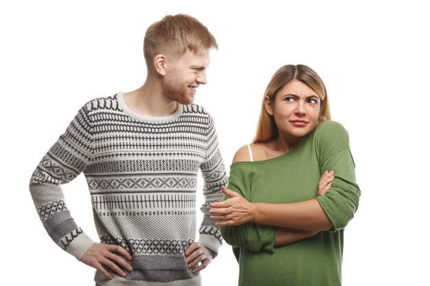 Handsome bearded guy dressed in sweater smiling and looking at attractive woman who standing in closed posture with arms crossed, feeling confused as she doesn't like or understand his silly joke